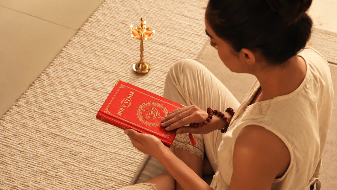 Žena sedí na tkané rohoži a čte červenou knihu Holy Vedas vedle mosazné ájurvédské lampy, pro Art of Vedas